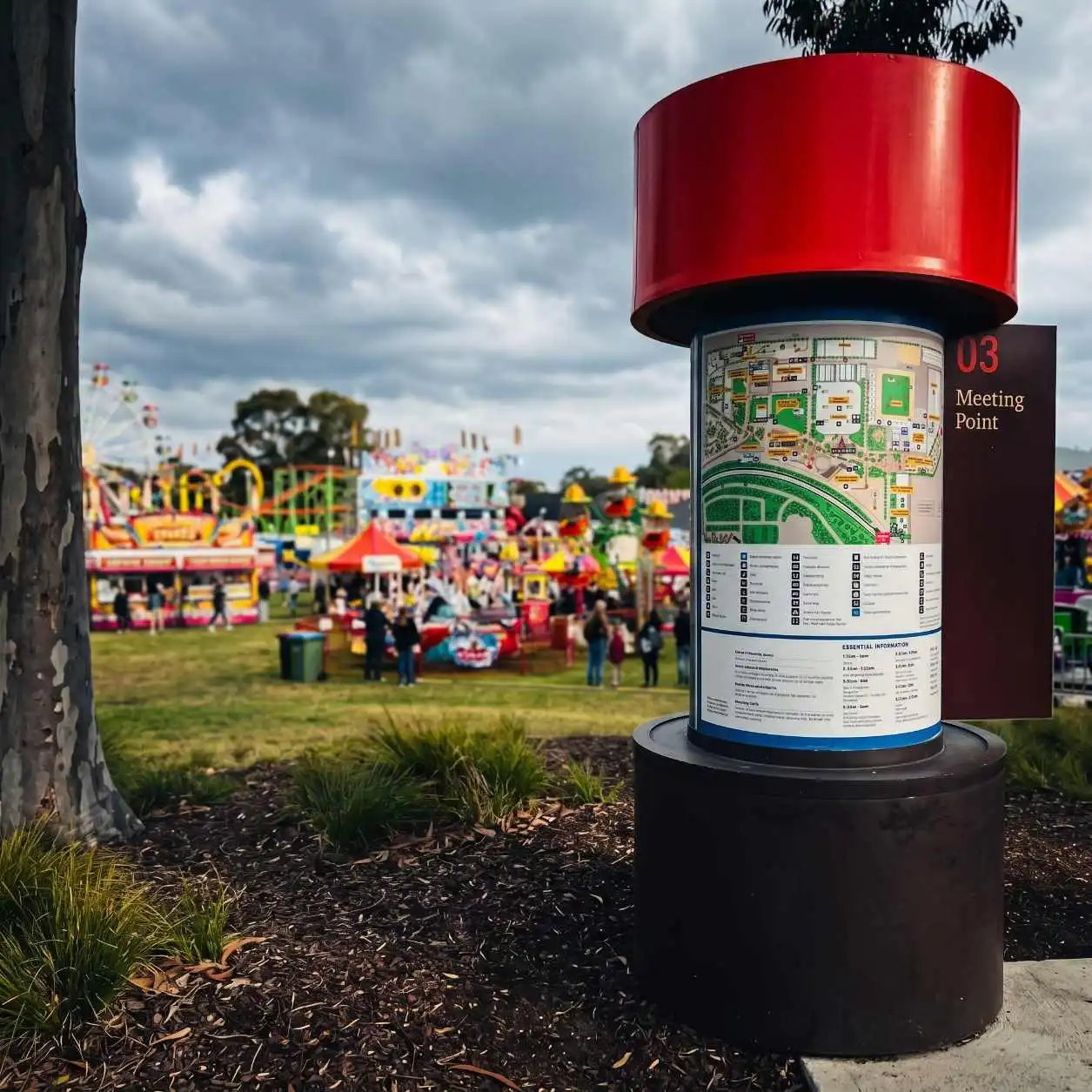 Map kiosk with a bright red cylindrical top at a festival; sign reads '03 Meeting Point' beside a detailed map.